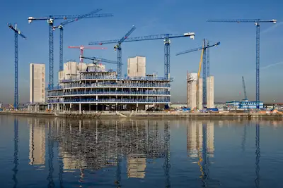 Große Baustelle mit Kränen und Rohbau neben einer Wasserfläche.
