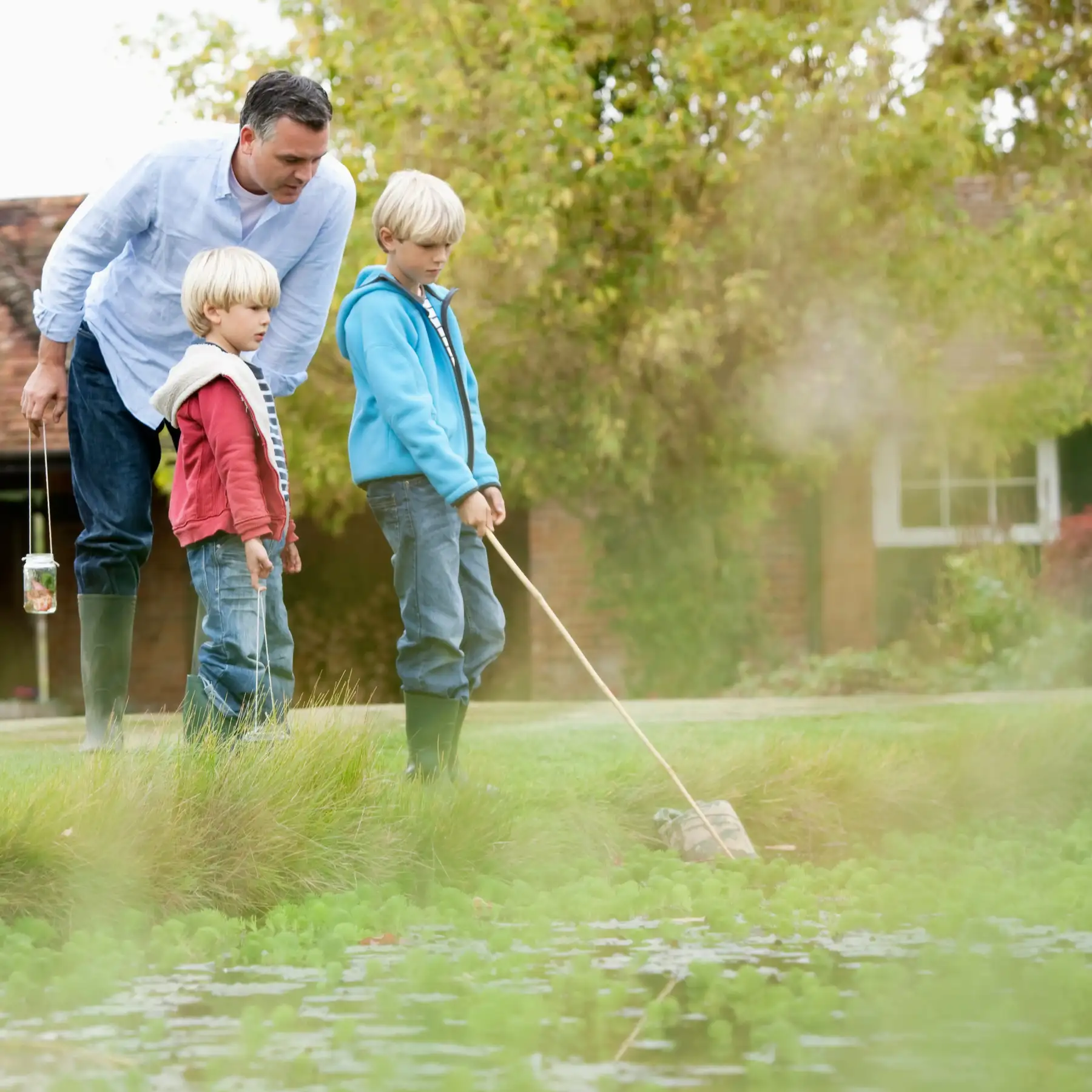 Vater begleitet zwei Kinder beim Kescherfang und Beobachten der Natur an einem Teich.
