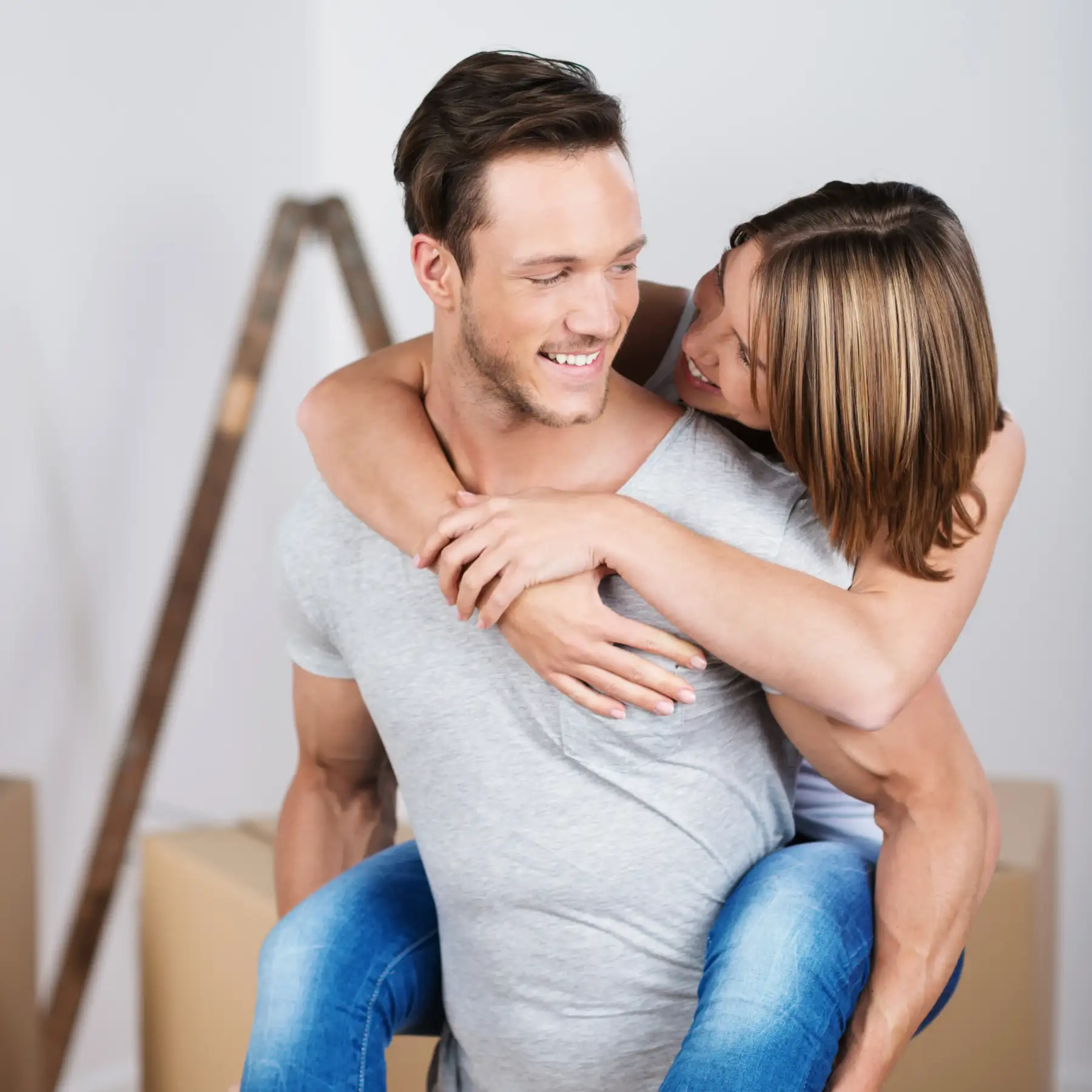 Frau umarmt ihren Partner von hinten; beide lachen in einer neuen Wohnung mit Umzugskartons.