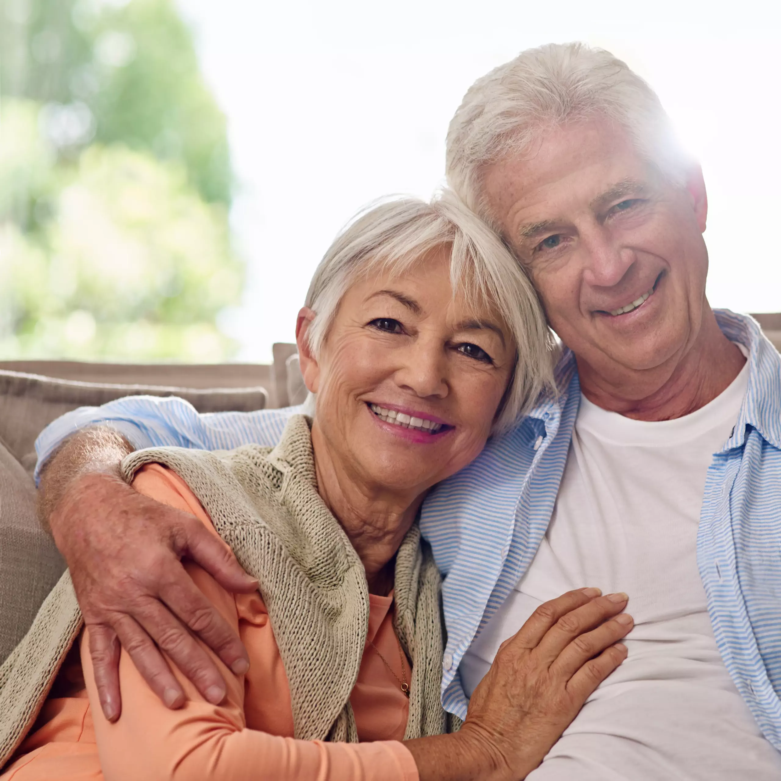 Älteres Paar umarmt sich lächelnd in einem hellen Wohnraum.